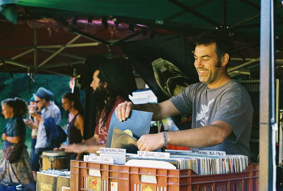 A smiling record seller sits beside vinyl records and a hand drum.
