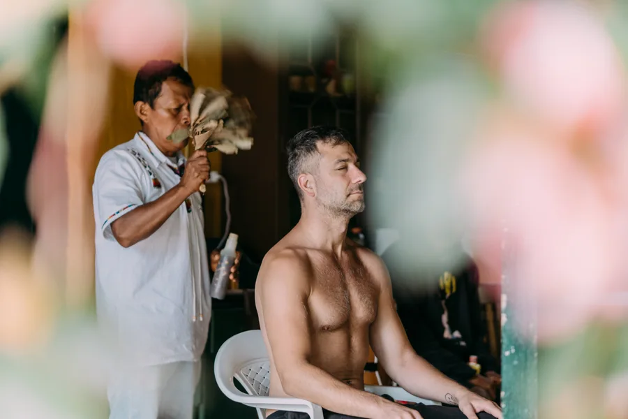 A seated man closes his eyes while another person holds feathers near him.